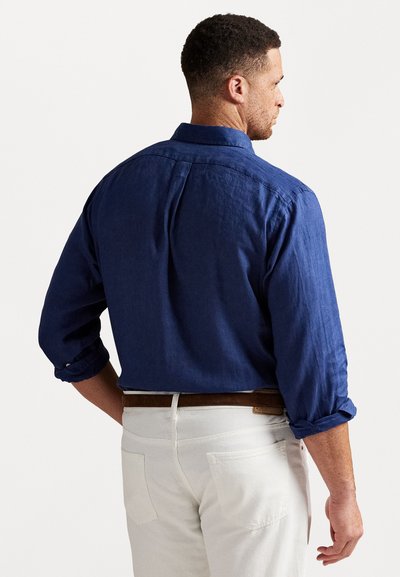 Marinblå skjorta med knappar och uppvikta ärmar, tillverkad av textil med struktur. Bakdelen har en yoke och plissering, tillsammans med krämfärgade byxor.