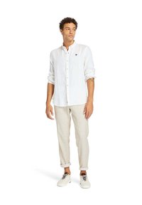 Camicia button-up bianca in lino con un piccolo logo, abbinata a pantaloni beige con risvolto e sneaker bianche. Design semplice e pulito con tessuto leggero.