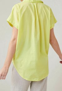 Korte mouwen shirt van heldergeel stof, met een rugplooitje en een gebogen zoom, gecombineerd met lichtgekleurde textuurbroek.