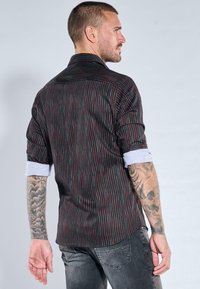 Schwarzes Hemd mit dünnen vertikalen roten und blauen Streifen, geknöpfter Kragen, lange Ärmel an den Bündchen aufgerollt, Rückansicht zeigt ein tailliertes Design.