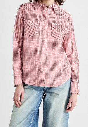 Kvinde iført rød-hvid ternet skjorte med lange ærmer og trykknapper, kombineret med lyseblå falmede jeans mod ensfarvet baggrund.