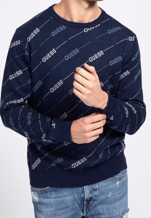 Hombre con un suéter azul marino que presenta un patrón diagonal repetido de texto "GUESS", combinado con jeans claros desgastados.