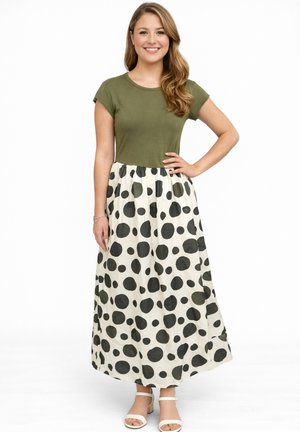 Kobieta stoi z ręką na biodrze, ubrana w oliwkowo zieloną bluzkę z krótkim rękawem oraz białą długą spódnicę w duże czarne kropki i białe sandały.