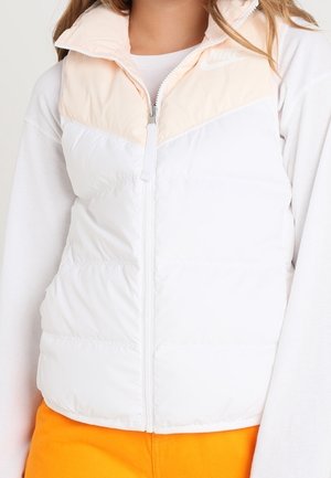 Woman wearing a white and light pink sleeveless puffer vest with a front zipper over a white long-sleeve shirt and bright orange pants.