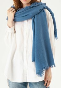Écharpe bleue en tissu doux, ornée de bords à franges, drapée de manière décontractée sur une chemise blanche à boutons, couvrant une partie du bras de la personne qui la porte.