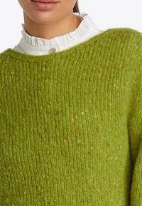 Groene gebreide trui van een zachte, gestructureerde wollen mix, met een brede halslijn en een geribd patroon, gedragen over een witte trui met ruches aan de kraag.
