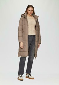 Gepolsterter langer Mantel in Taupe, mit Kapuze, Seitentaschen und Reißverschluss. Getragen über einem beigen Strickpullover und dunklen Jeans mit gemusterten Sneakers.