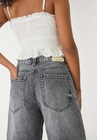 Haut blanc à smocks avec un ourlet volanté, associé à un jean en denim gris taille haute avec poches arrière et une étiquette en cuir près de la taille.