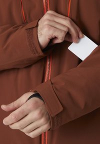 Hnědá nepromokavá bunda s oranžovým zipem, se zápěstím na suchý zip. Ruka sahá pro kartu z kapsy na rukávu.