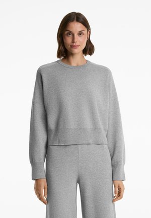 Vrouw in een lichtgrijze lange mouwen crewneck trui en bijpassende wijde broek, staand tegen een effen witte achtergrond.