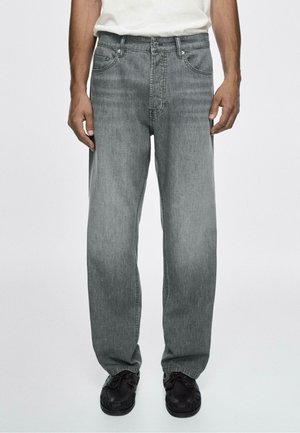 Man draagt losse grijze denim jeans en zwarte casual schoenen, staand tegen een eenvoudige lichte achtergrond.