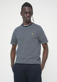 Μπλούζα με ριγέ ναυτικό και άσπρο χρώμα σε απαλό ύφασμα, με στρογγυλή λαιμόκοψη και κίτρινο λογότυπο πουλιού στο αριστερό στήθος. Κοντά μανίκια, άνετη γραμμή.