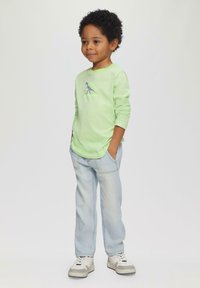 Helle grüne Langarmbluse mit einem Dinosaurierdruck, kombiniert mit hellblauen Jeans und weißen Turnschuhen. Die Hände des Kindes sind in den Taschen.