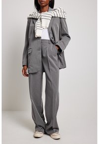 Costume gris sur mesure avec une veste ample et un pantalon à jambes larges. Pull en maille blanche drapé sur les épaules, associé à des chaussures décontractées.