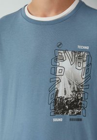 Blaues Baumwoll-T-Shirt mit Rundhalsausschnitt, das einen schwarz-weißen Grafikdruck einer Konzertszene zeigt, umgeben von fettem schwarzen Text.