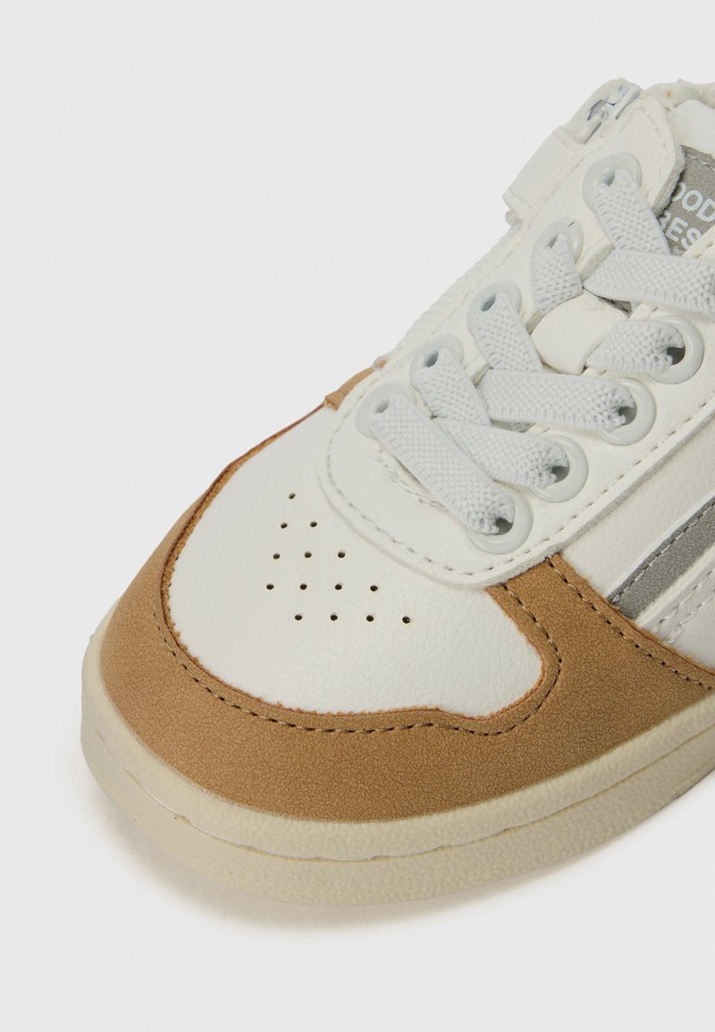 Sneaker in pelle con punta bianca, accenti in suede beige, strisce grigie e suola in gomma testurizzata. I lacci sono di un grigio chiaro, con dettagli perforati.