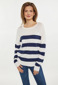 Pulover cu mâneci lungi, dungi în alb și bleumarin, cu o decolteu larg și manșete îndoite, purtat cu blugi ajustați, albaștri.