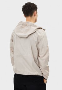 Ljusbeige vindjacka med huva, elastiska muddar och resår i fållen. Tillverkad av slät, lättviktig tyg. Enkel och funktionell design.