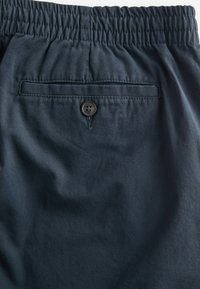 Pantalones cortos azul marino con cinturilla elástica, que cuentan con un bolsillo trasero asegurado por un botón negro. Tejido suave con un ligero brillo.