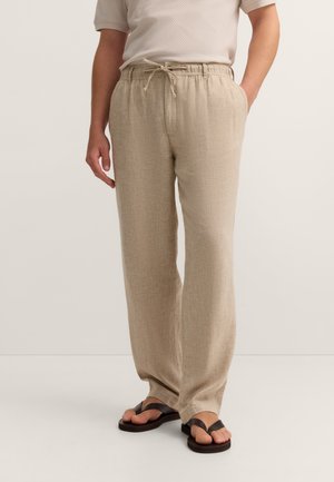 Mann trägt beige Leinenlatzhose mit Tunnelzug, weißes kurzärmeliges Hemd und schwarze Flip-Flops und steht vor einem einfarbigen Hintergrund.