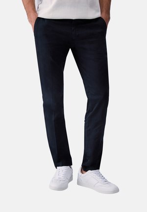 Uomo che indossa pantaloni neri slim fit e scarpe da ginnastica bianche, mani in tasca, in piedi davanti a uno sfondo semplice.
