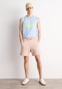 Chemise sans manches bleu clair avec un motif jaune, short rose avec un logo bleu, casquette beige, chaussures blanches avec des semelles beiges. Tenue décontractée d'été.