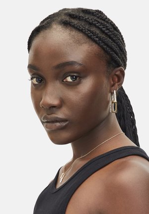 Silberne geometrische Ohrringe mit rechteckigem Design; getragen von einer Person mit geflochtenem Haar, kombiniert mit einer schlichten goldenen Halskette und einem schwarzen Oberteil.