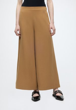 Kvinde iført camel-farvede bukser med høj talje og vide ben, kombineret med sorte lakflade sko med spændelukning og en sort ærmeløs top.