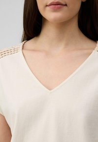 Haut crème à col en V avec des manches courtes, présentant un détail en crochet texturé le long des épaules. Tissu en coton doux, coupe décontractée.