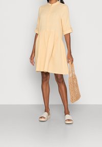 Light yellow button-up dress made of textured fabric, with three-quarter sleeves and a tiered skirt, paired with beige slide sandals and a woven bag.