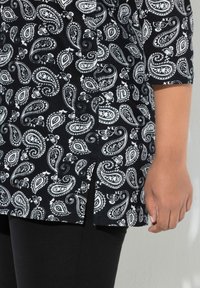 Schwarze Tunika mit weißem Paisleymuster, mit kurzen Ärmeln und Seitenschlitzen. Schwarze Leggings darunter sichtbar. Weiche Stofftextur.