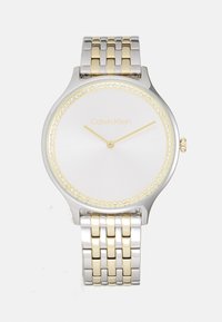 Reloj en tonos plateado y dorado con caja redonda de acero inoxidable, bisel de cristal, esfera blanca minimalista y pulsera de metal vinculada.