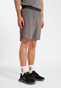 Gråa sportshorts med resår i midjan, med en orange varumärkeslogotyp och en chevron-mönster på sidan. Bärs med svarta skor och vita strumpor.
