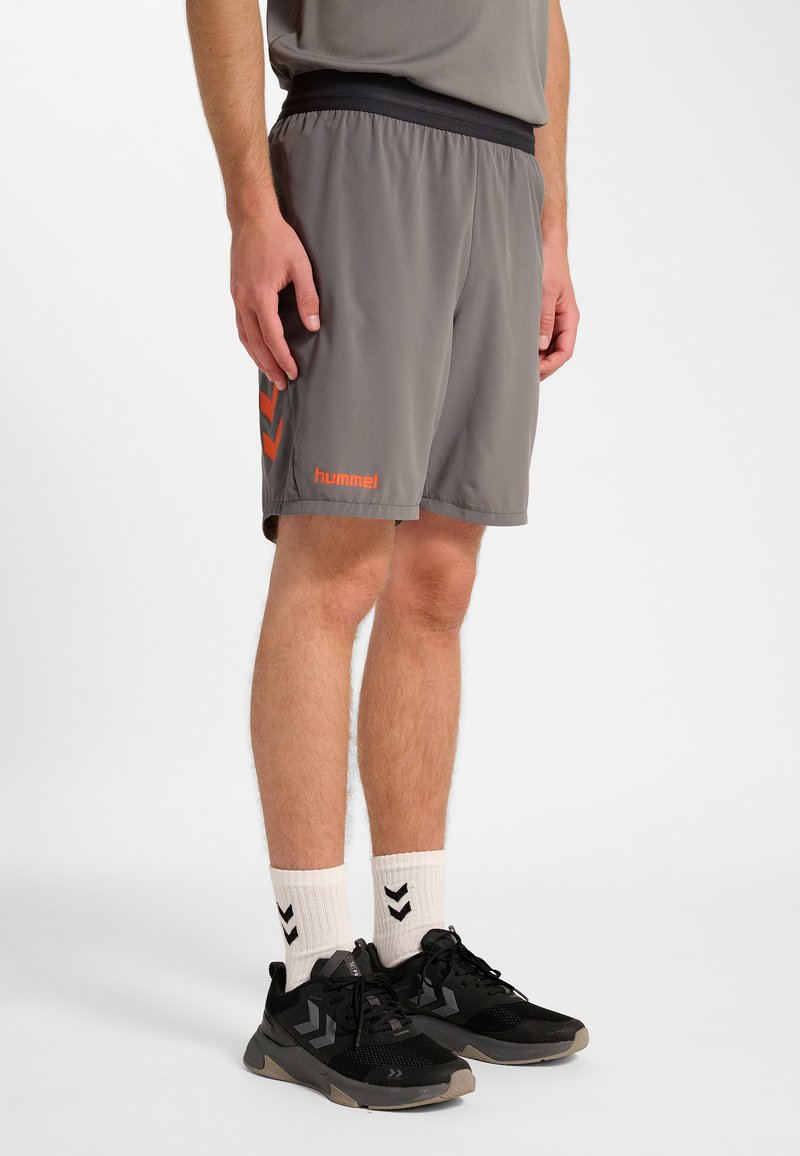 Gråa sportshorts med resår i midjan, med en orange varumärkeslogotyp och en chevron-mönster på sidan. Bärs med svarta skor och vita strumpor.