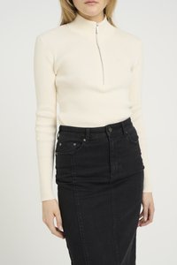 Top a maniche lunghe a coste crema con colletto alto e cerniera argentata, abbinato a una gonna di jeans nera con tasche frontali e vestibilità slim.