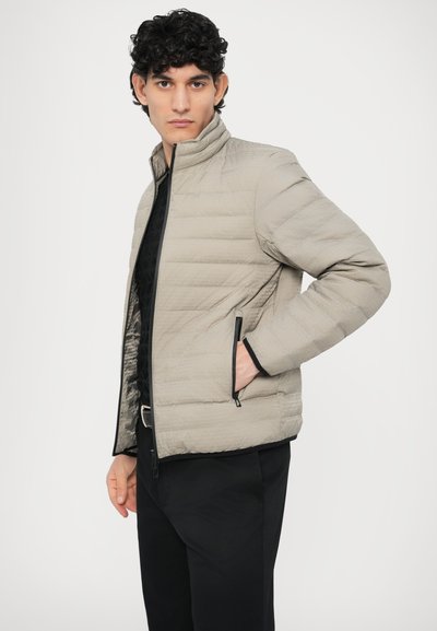 Svetlo siva puhasta jakna z visokim ovratnikom, horizontalnim šivom in črnimi zadrgami. Nošena preko črne majice z vzorcem.