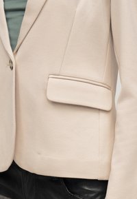 Blazer beige sur mesure avec poche à rabat porté sur un haut vert et associé à un pantalon en cuir noir.
