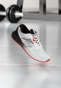 Άσπρο, μαύρο και κόκκινο παπούτσι τρεξίματος Reebok που αιωρείται πάνω από γυαλιστερό δάπεδο με θολά βάρη γυμναστηρίου στο παρασκήνιο.