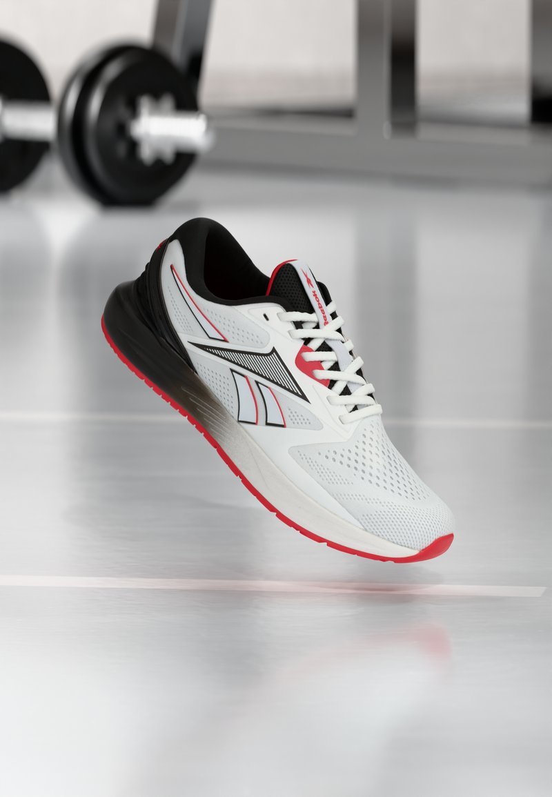 Άσπρο, μαύρο και κόκκινο παπούτσι τρεξίματος Reebok που αιωρείται πάνω από γυαλιστερό δάπεδο με θολά βάρη γυμναστηρίου στο παρασκήνιο.