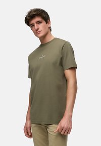 Camiseta de algodón verde oliva con cuello redondo, mangas cortas y un pequeño logo blanco centrado en el pecho. Textura suave, corte casual.