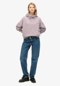 Jasnofioletowa bluza z kapturem, z sznurkiem, kieszenią kangurką i subtelnym logo; zestawiona z niebieskimi jeansami o wysokim stanie i czarnymi butami.