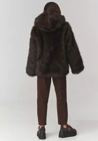 Brązowa kurtka z futra sztucznego z kapturem, w oversize'owym fasonie i długimi rękawami, w połączeniu z brązowymi spodniami i masywnymi czarnymi butami, widok z tyłu.