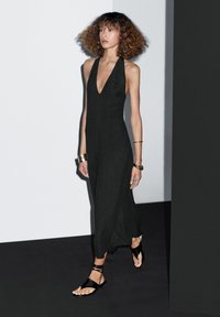 Black sleeveless dress with a deep V-neck and flowing shape. Model wears black strappy sandals, with a smooth, lightweight fabric texture.