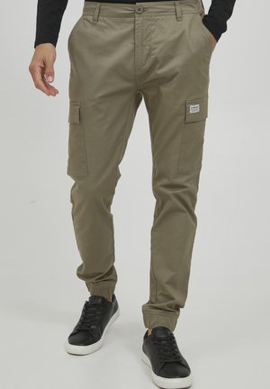 Khaki bojówki z regulowanymi mankietami u kostek, kieszeniami po bokach oraz małą naszywką z logo. W zestawie z czarnymi sneakersami z białą podeszwą.