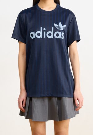 Femme portant un t-shirt Adidas bleu foncé avec un logo bleu clair et une jupe plissée grise, debout devant un fond uni.