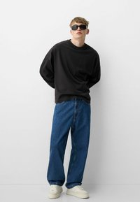 Čierna voľná mikina, modré voľné džínsy a biele tenisky. Model s čiernymi slnečnými okuliarmi stojí na jednotnom pozadí.