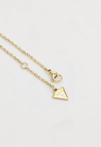 Gouden ketting met een getextureerde driehoekige hanger waarop het woord "GUESS" staat. De ketting heeft een karabijnsluiting.
