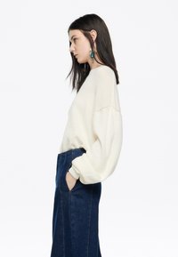 Młoda kobieta z długimi ciemnymi włosami nosi biały, oversize'owy sweter oraz spódnicę z wysokim stanem w ciemnoniebieskim denimu, z rękami w kieszeniach, zwrócona w lewo.