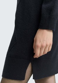 Hand rustend tegen een zwarte gebreide jurk met geribbelde zoom en zijslit, gedragen over doorschijnende zwarte panty’s.