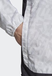 Leichter Windbreaker mit einem grau-weißen Streifenmuster, elastischen Bündchen und einem schwarzen unteren Bereich. Die Tasche ist an der Seite sichtbar.